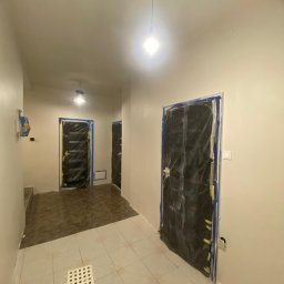 Twoja Ekipa Remontowa Konrad Lewicki - Korytarz w trakcie prac wykończeniowych: świeżo pomalowane ściany, zabezpieczone folią drzwi, widoczny stołek malarski i surowe oświetlenie.