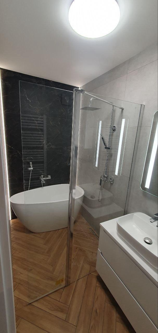 Nowoczesna łazienka z wolnostojącą wanną na tle czarnej ściany z marmurowym wzorem, kabiną prysznicową ze szkła i drewnopodobną podłogą w jodełkę.