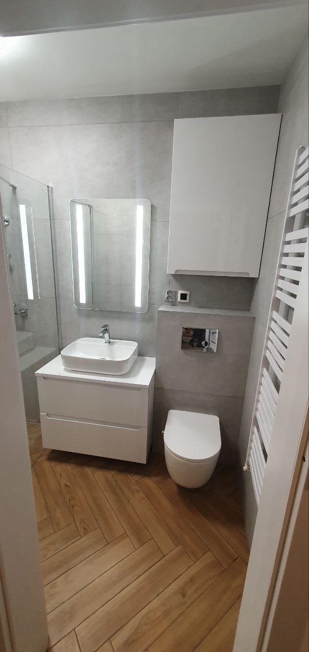 Nowoczesna łazienka z drewnianą podłogą w jodełkę, szarą płytką i białymi meblami: umywalką, szafką i grzejnikiem drabinkowym. Lustro z podświetleniem LED.