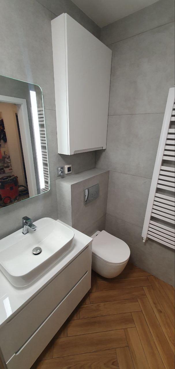 Nowoczesna łazienka z szarymi płytkami, białą szafką pod umywalką, lustrem z podświetleniem LED i drewnianą podłogą ułożoną w jodełkę. Minimalistyczny design.