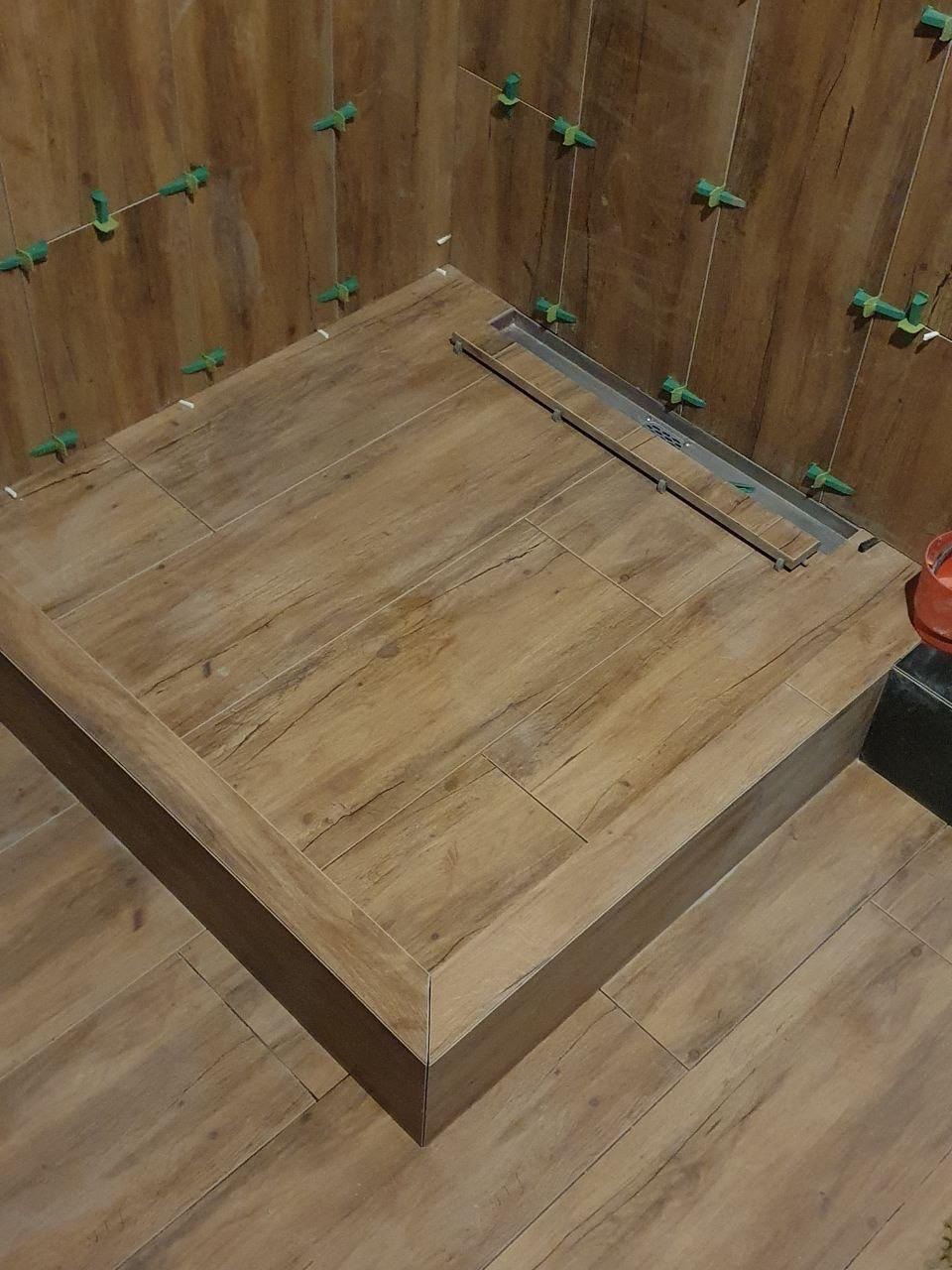 Wykończony brodzik z drewnopodobnych płytek, z zamontowanym odpływem liniowym i zielonymi krzyżykami dystansowymi. Widoczne fugi i precyzyjne cięcia płytek.