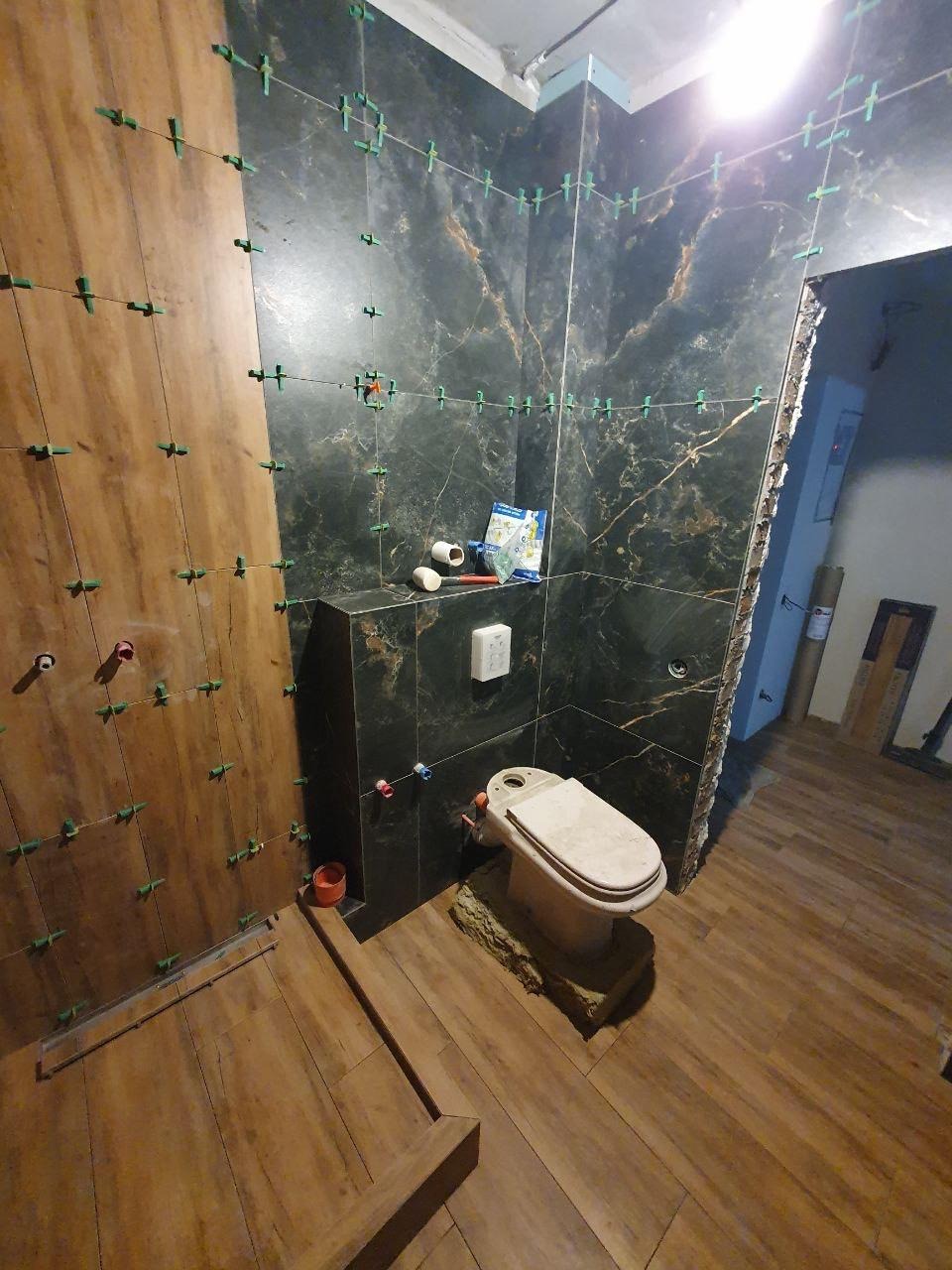 Układanie płytek imitujących drewno na podłodze i czarnych płytek z marmurowym wzorem na ścianach łazienki, z użyciem systemu poziomowania płytek.