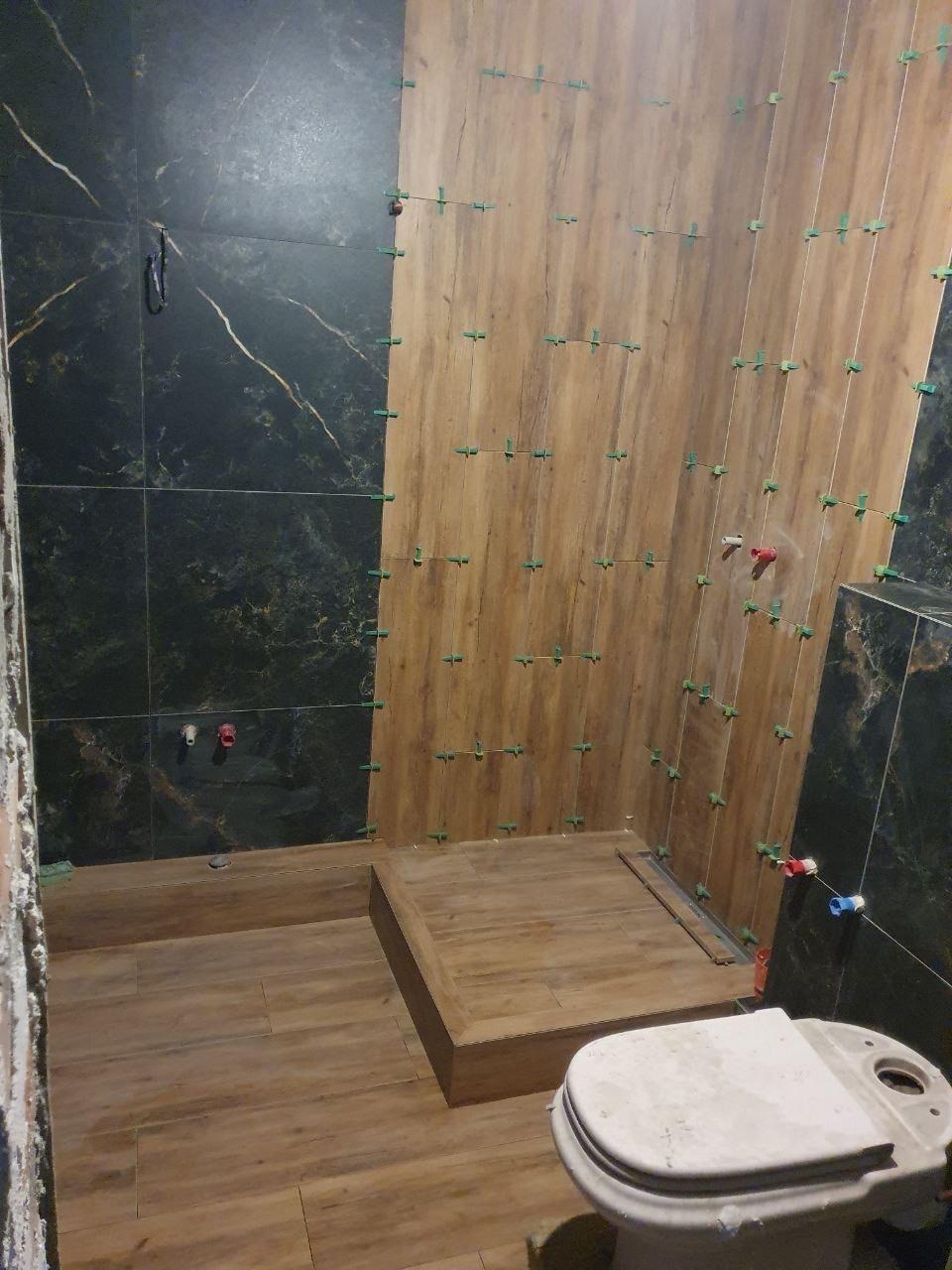 Układanie płytek imitujących drewno i czarnego marmuru w łazience. Widoczne krzyżyki dystansowe i fragment toalety. Prace w toku, surowy stan.
