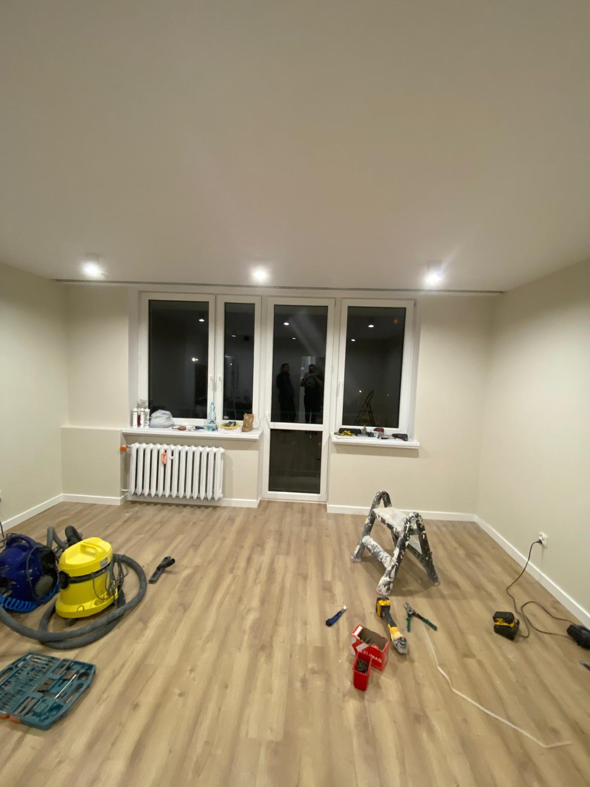 Jasne wnętrze po remoncie: świeżo pomalowane ściany, nowe panele podłogowe, okno balkonowe. Widoczne narzędzia i drabina malarska.