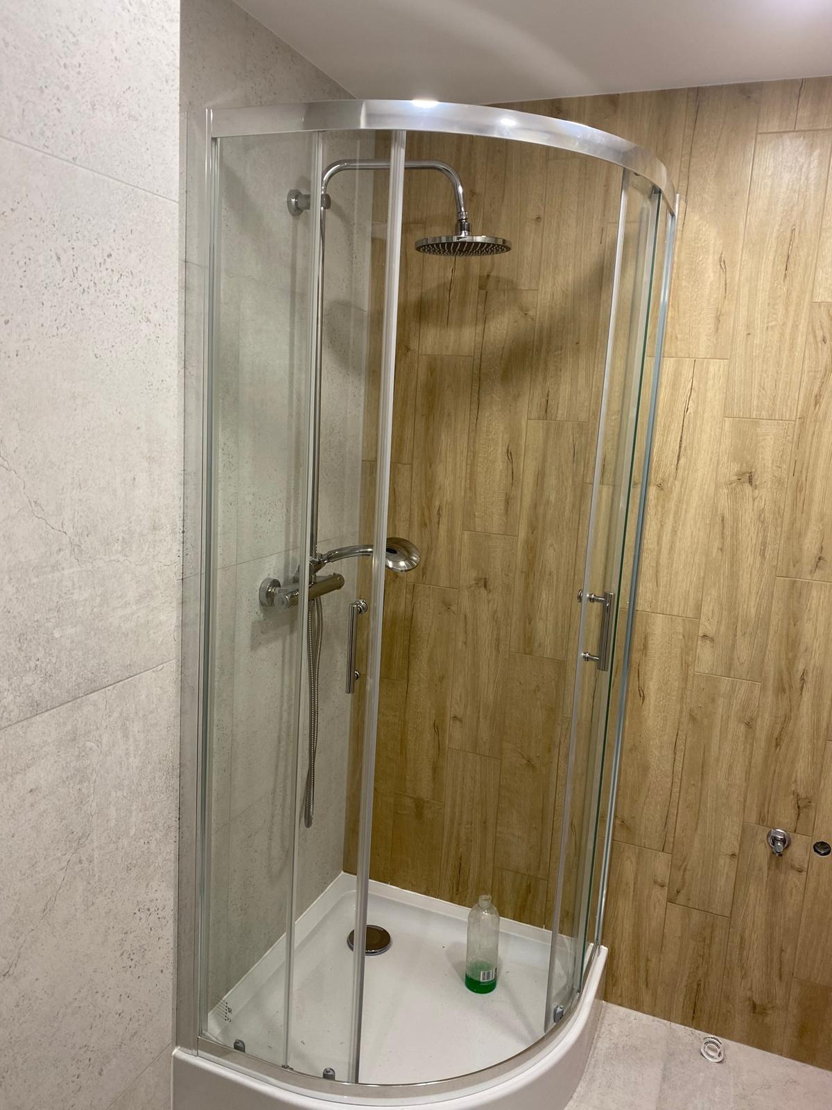 Nowoczesna kabina prysznicowa z chromowanymi elementami i deszczownicą, otoczona płytkami imitującymi drewno i kamień. Widoczna butelka z płynem na brodziku.