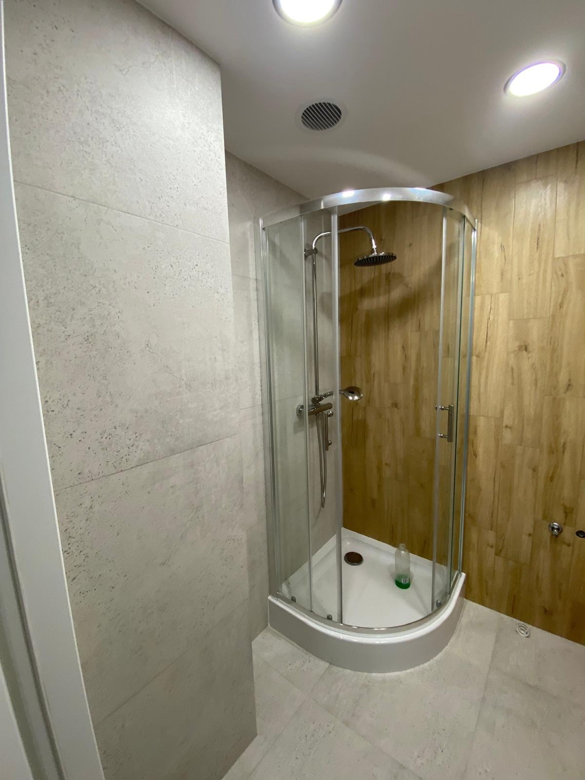 Nowoczesna kabina prysznicowa z chromowanymi elementami, wykończona płytkami imitującymi drewno i kamień, z widoczną butelką kosmetyku wewnątrz.