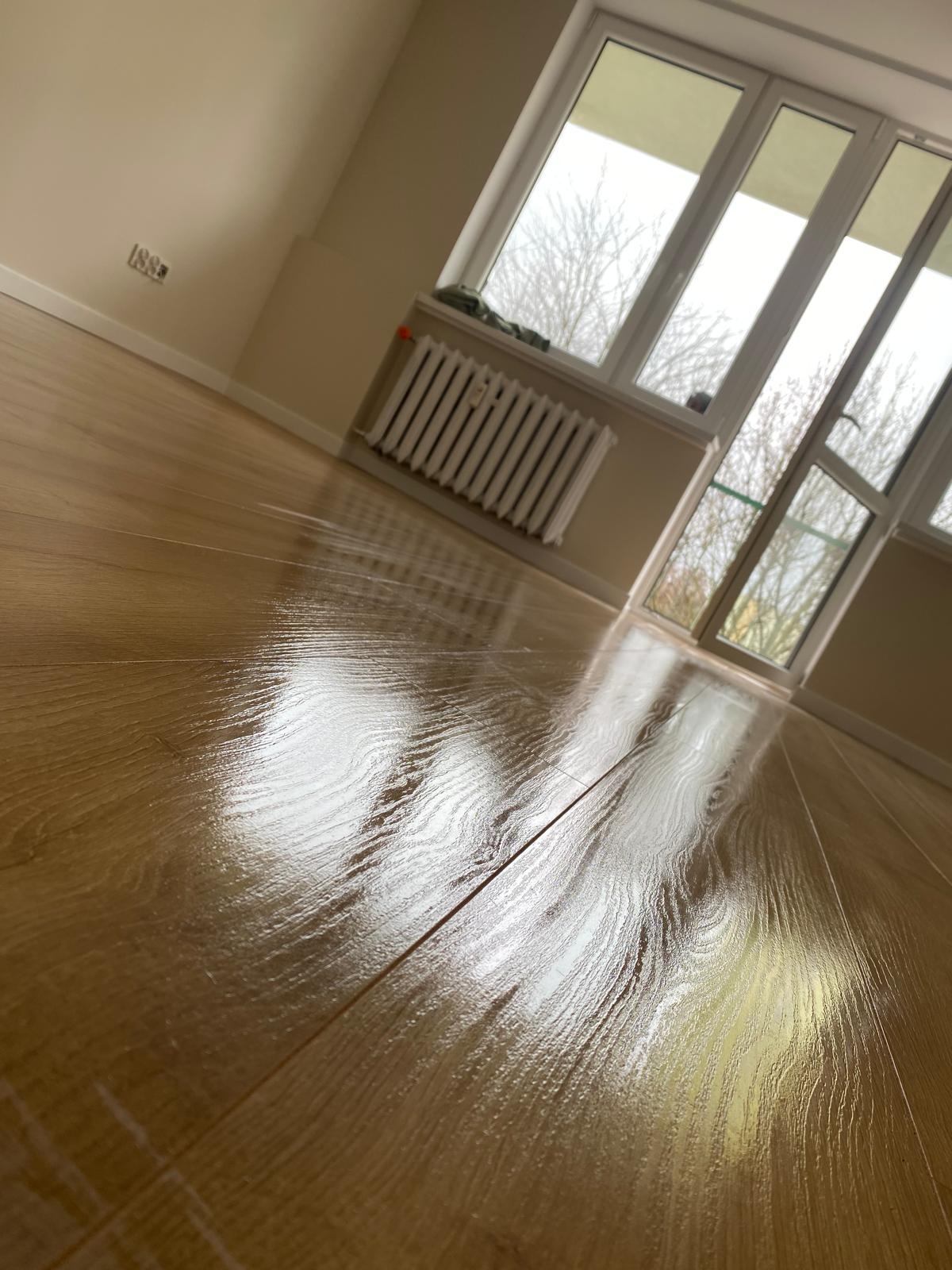 Błyszcząca, drewniana podłoga po renowacji w jasnym pokoju z oknem i grzejnikiem. Widoczne detale struktury drewna i odbicia światła.
