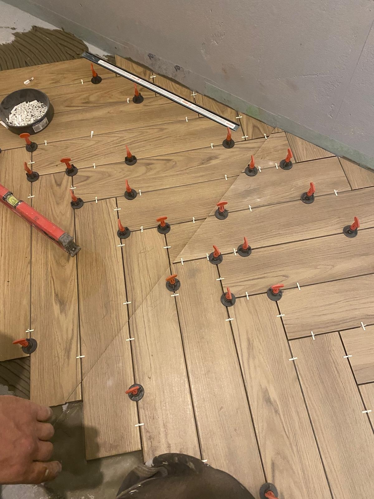 Układanie parkietu w jodełkę z użyciem systemu poziomowania płytek. Widoczny poziomica, wiadro z krzyżykami dystansowymi i fragment dłoni glazurnika.