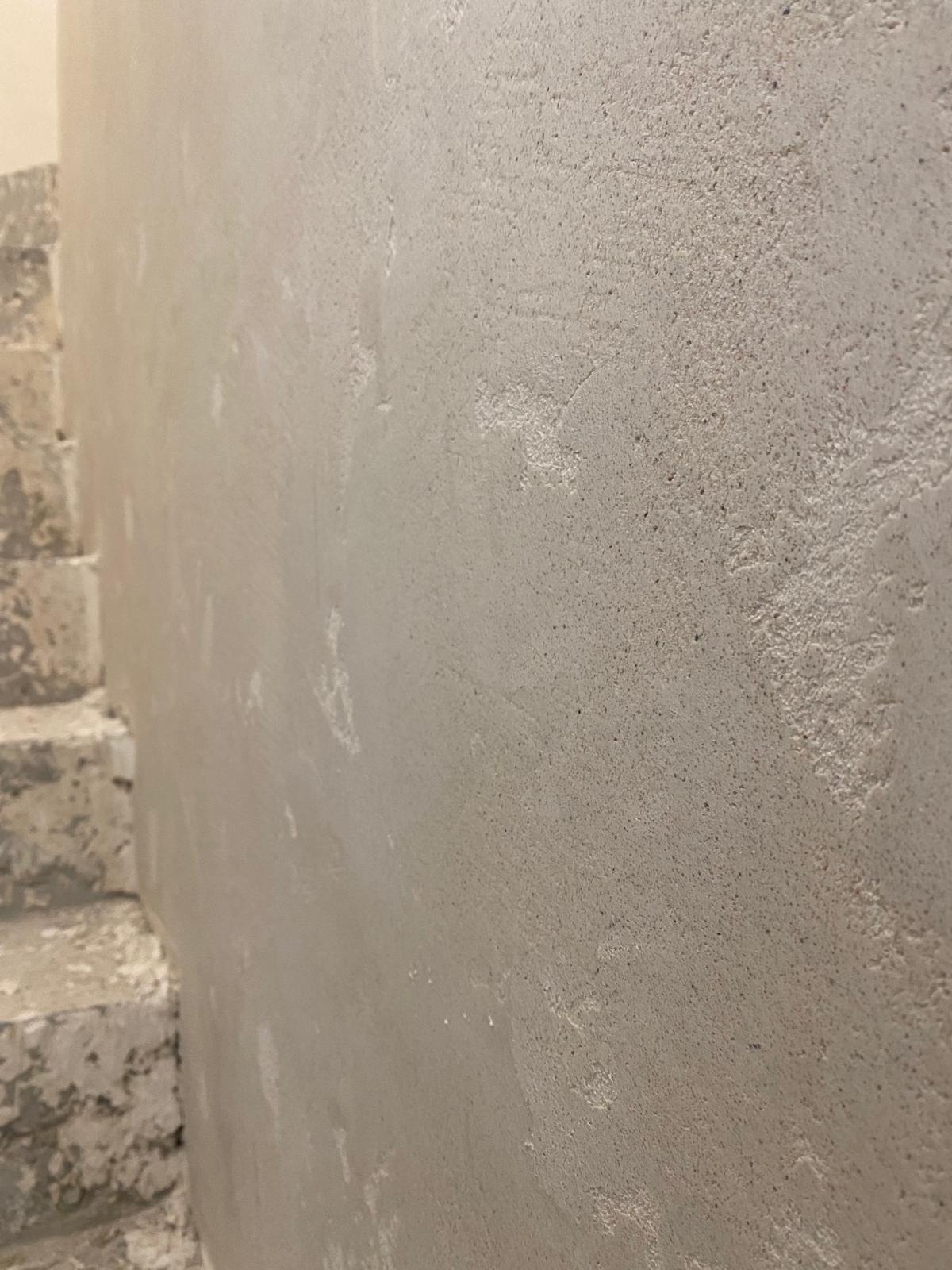 Szara, chropowata ściana po szpachlowaniu, widoczne nierówności i faktura. Fragment schodów z boku kadru. Neutralne oświetlenie.