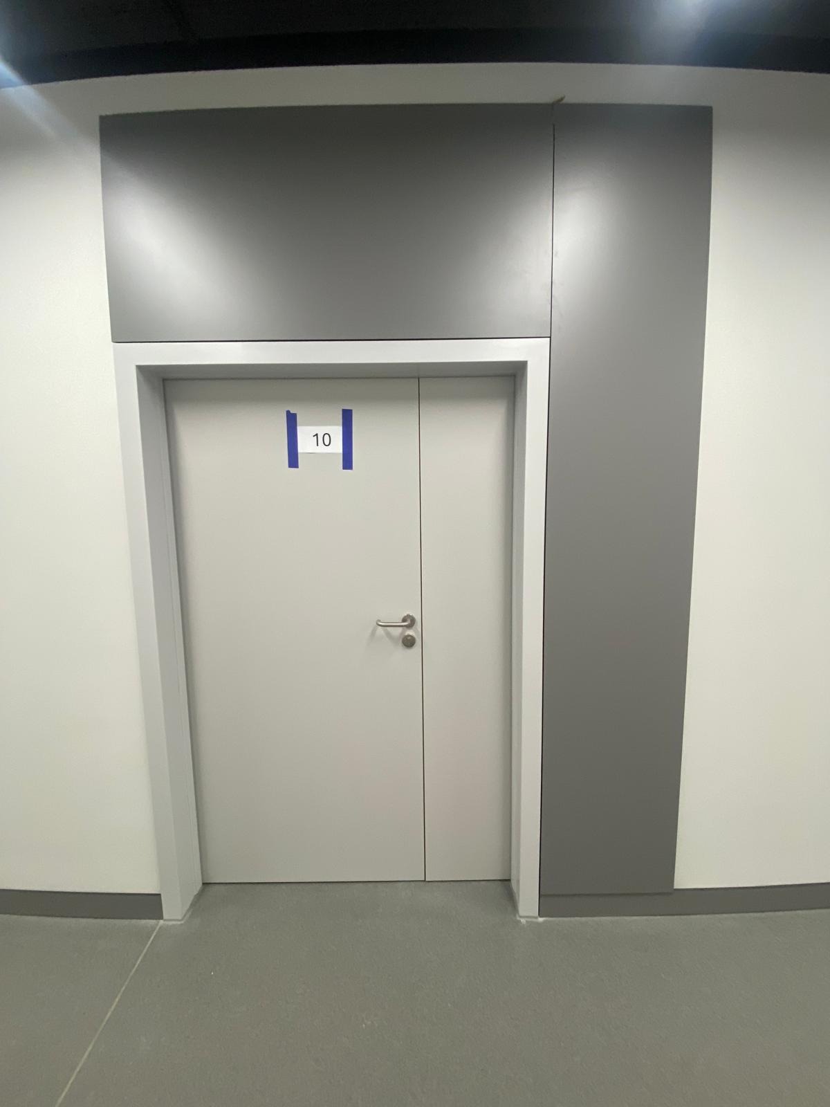 Białe drzwi z numerem 10, otoczone szarą ramą i panelami ściennymi w odcieniach szarości, na tle jasnej ściany i szarej podłogi. Minimalistyczny design wnętrza.