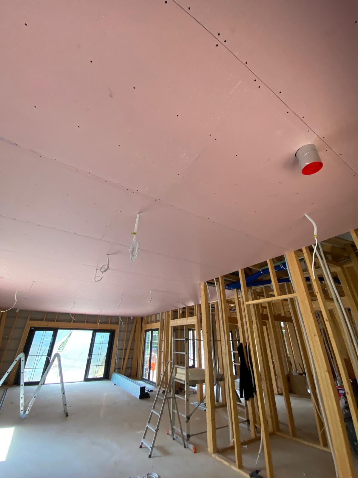 Montaż płyt karton-gips na suficie poddasza z widoczną konstrukcją drewnianą, instalacją i drabinami. Sufit w kolorze różowym.