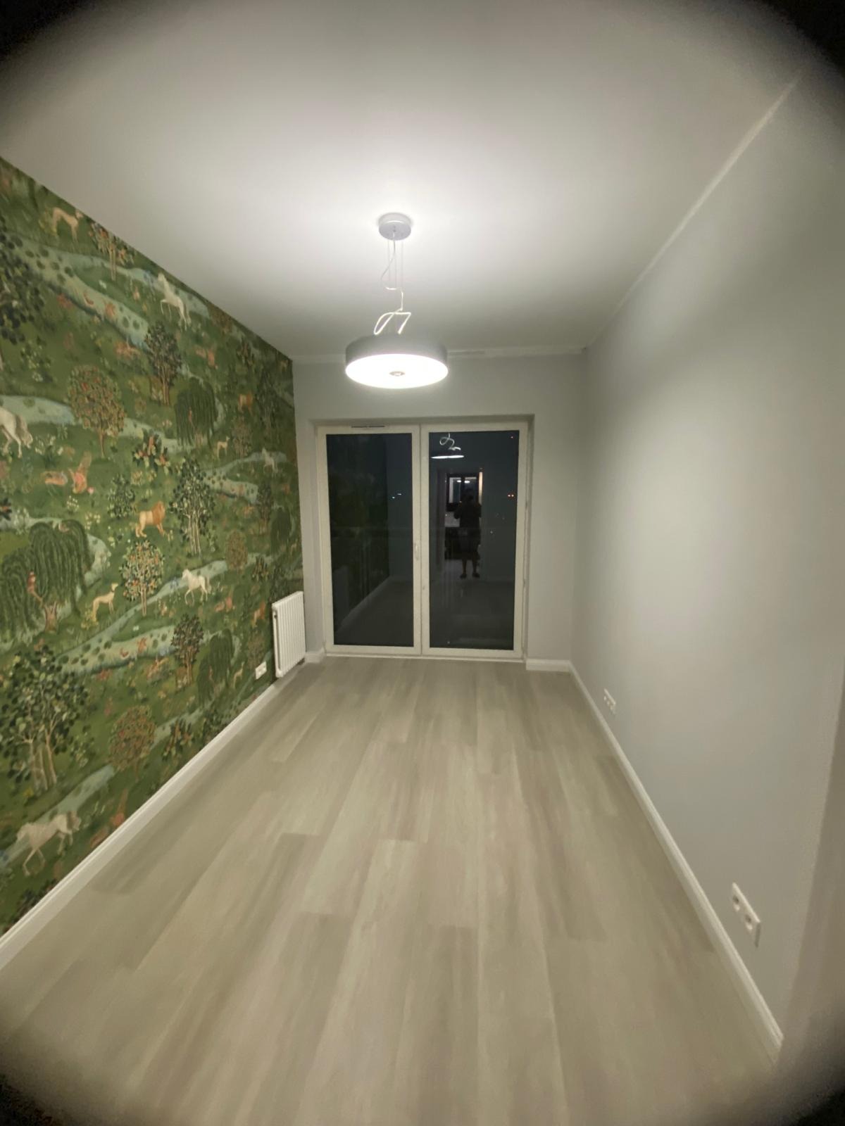 Pokój po remoncie: jedna ściana z tapetą w leśny motyw, druga pomalowana na szaro, jasna podłoga, białe listwy przypodłogowe i sufit, nowoczesna lampa.