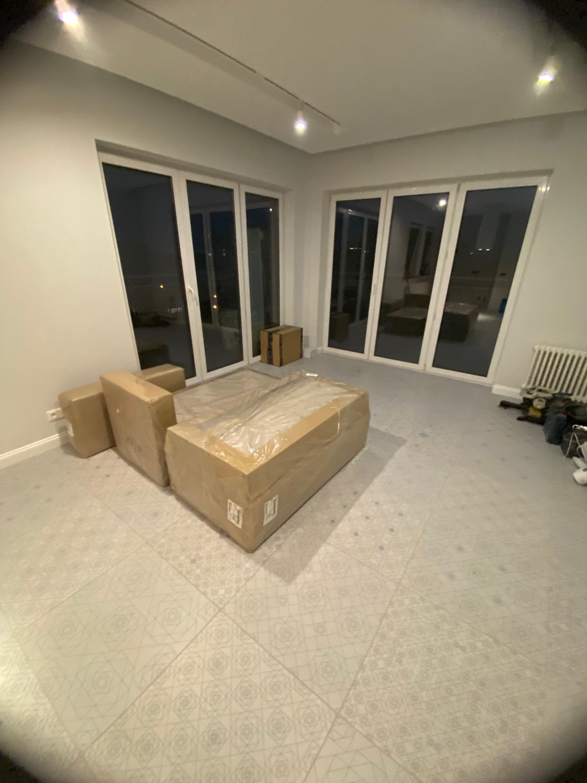 Nowe meble w kartonach na wzorzystej podłodze w jasnym pokoju z dużymi oknami. Przy grzejniku leżą narzędzia.