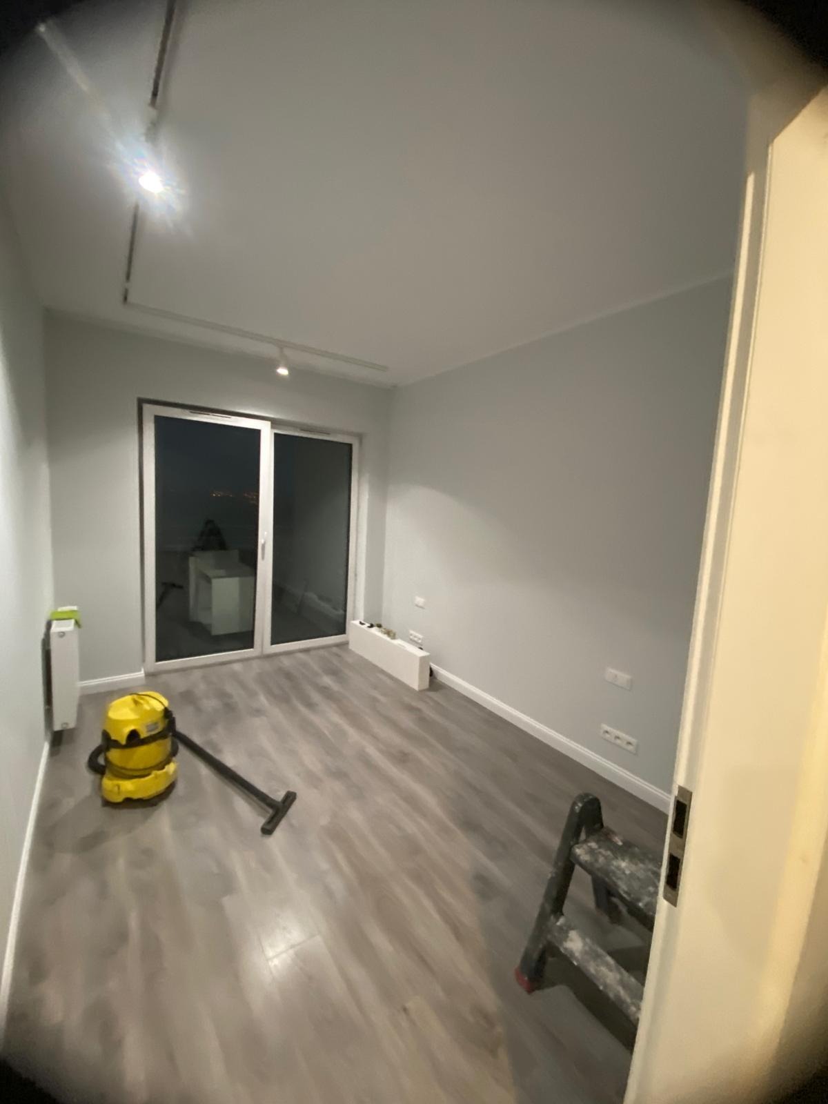 Pomalowane na szaro wnętrze pokoju z panelami, widoczny odkurzacz i drabina malarska. Świeżo wykończone ściany i podłoga po remoncie.