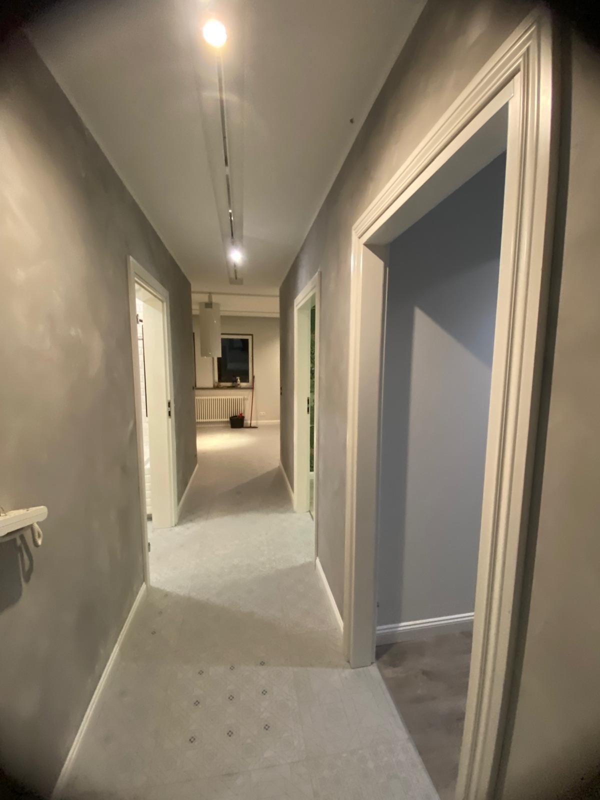 Korytarz po remoncie: szare ściany z efektem dekoracyjnym, białe futryny drzwi, wzorzysta podłoga i oświetlenie sufitowe. Widok w głąb mieszkania.