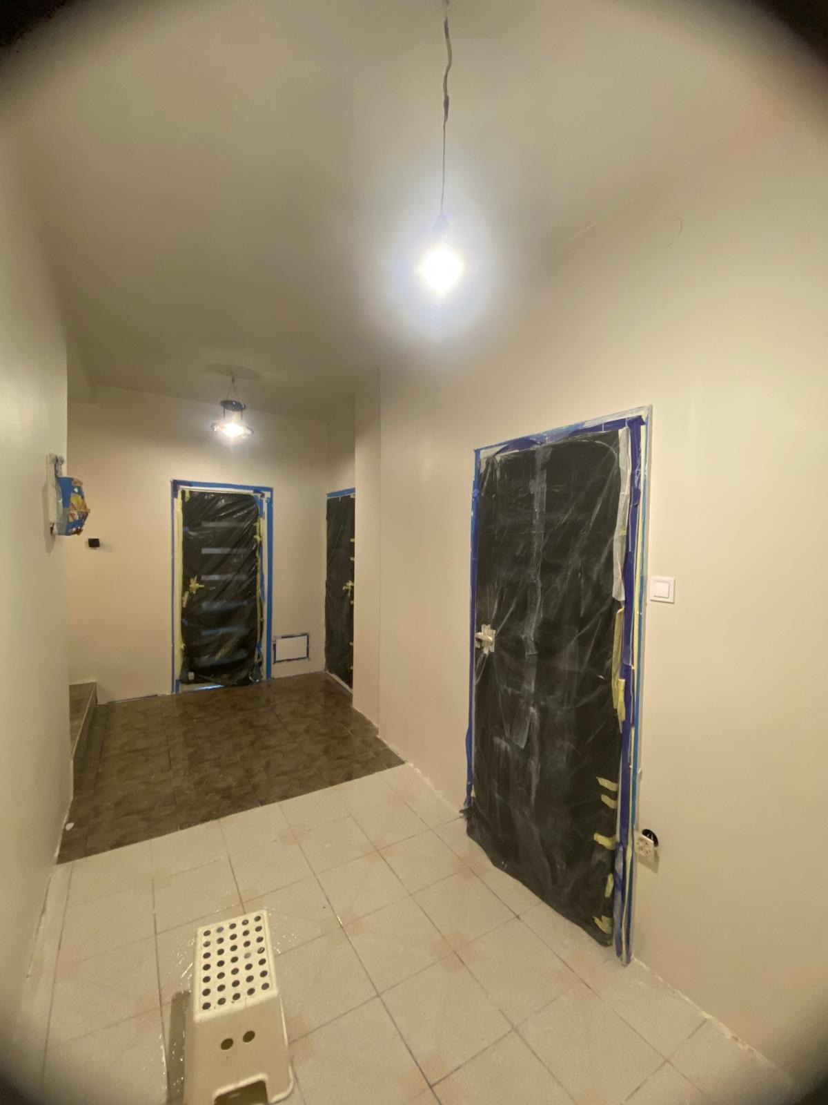 Korytarz w trakcie prac wykończeniowych: świeżo pomalowane ściany, zabezpieczone folią drzwi, widoczny stołek malarski i surowe oświetlenie.
