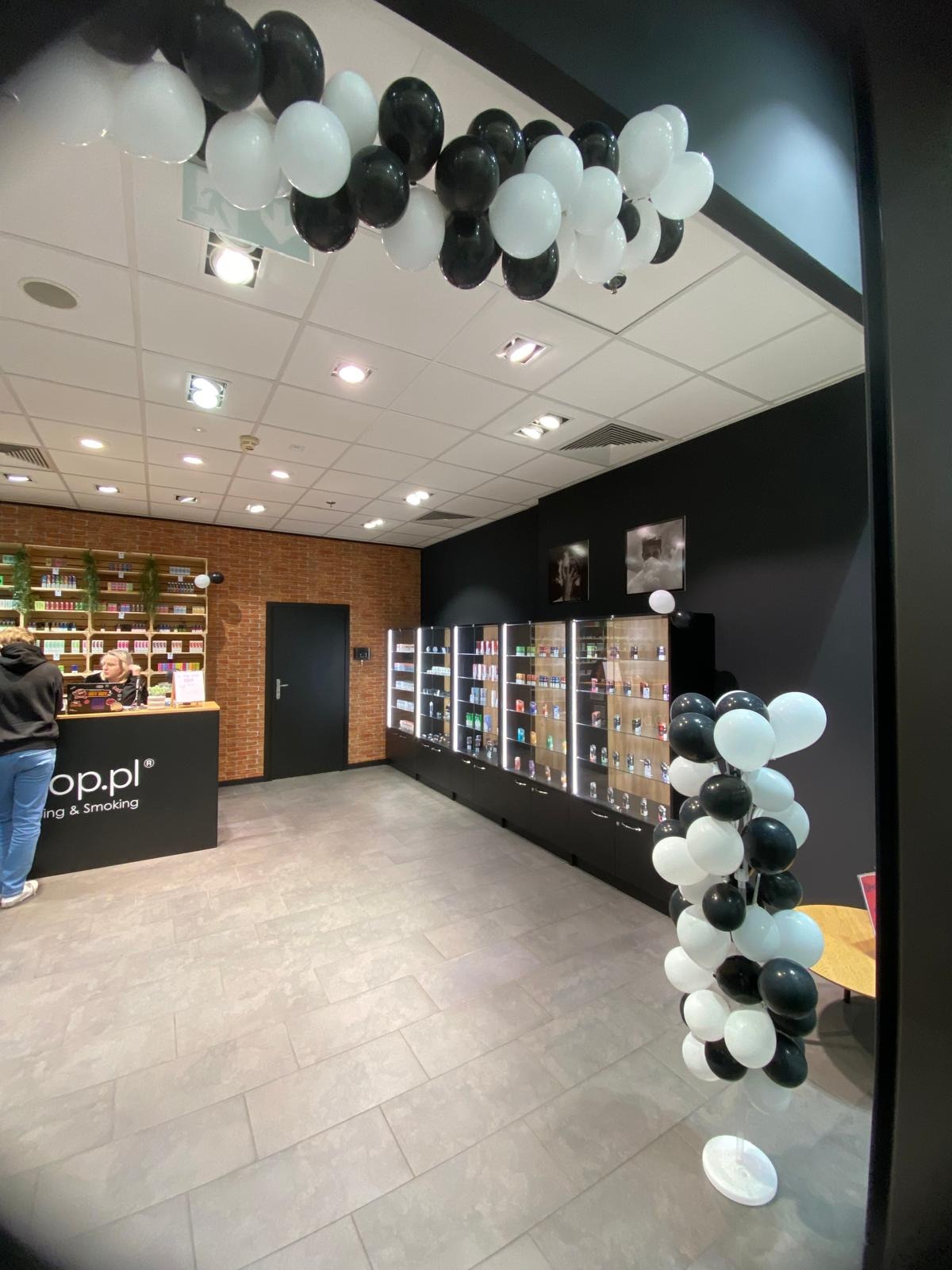 Nowoczesne wnętrze sklepu z ceglaną ścianą i czarnymi meblami, udekorowane białymi i czarnymi balonami. Widoczne regały z produktami i lada obsługi klienta.