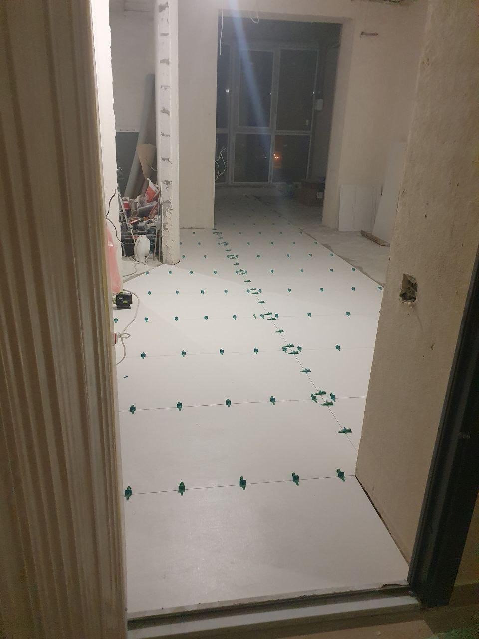 Biała podłoga z zielonymi elementami poziomującymi, przygotowana do dalszych prac wykończeniowych w remontowanym wnętrzu. Widok z perspektywy wejścia.