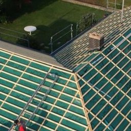 Dekarz Cieśla Stefan Kozanowski - Widok z góry na dach domu w trakcie krycia z zieloną membraną i drewnianą łatą, widoczne rusztowania i komin z cegły.