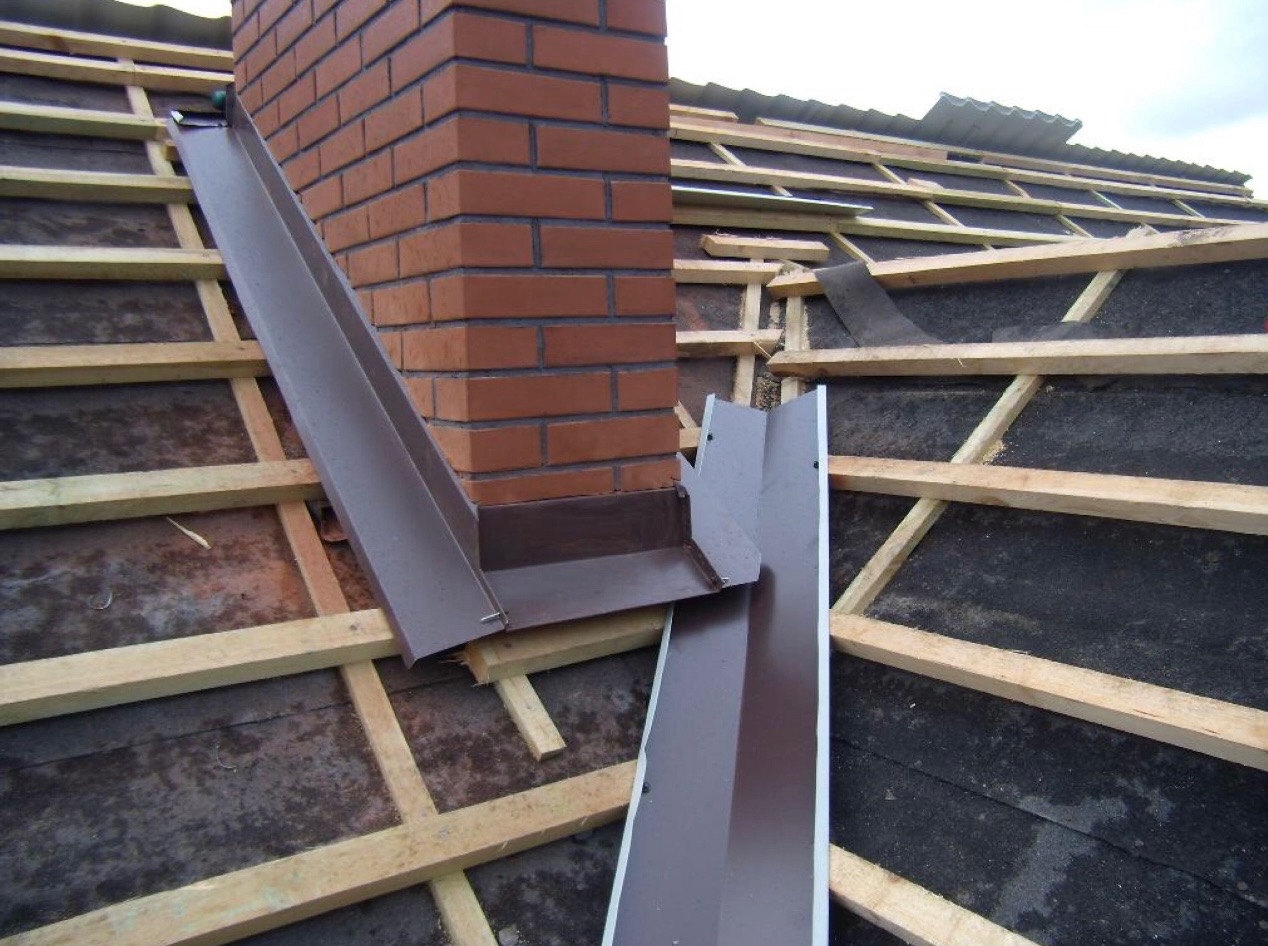 Montaż brązowych, metalowych elementów wykończeniowych dachu wokół komina z czerwonej cegły, widoczne drewniane krokwie i czarna membrana dachowa.