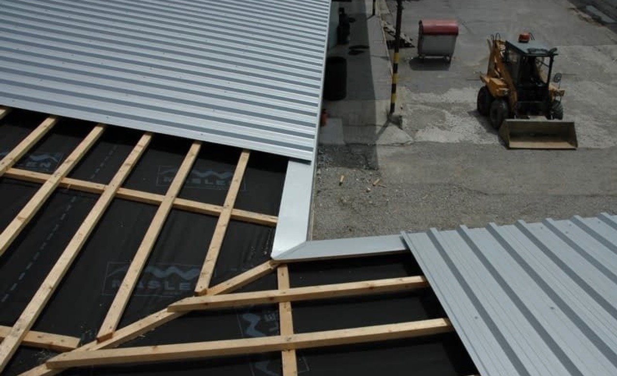 Widok z góry na częściowo pokryty blachą trapezową dach z widoczną drewnianą konstrukcją i czarną membraną, mała koparka w tle.