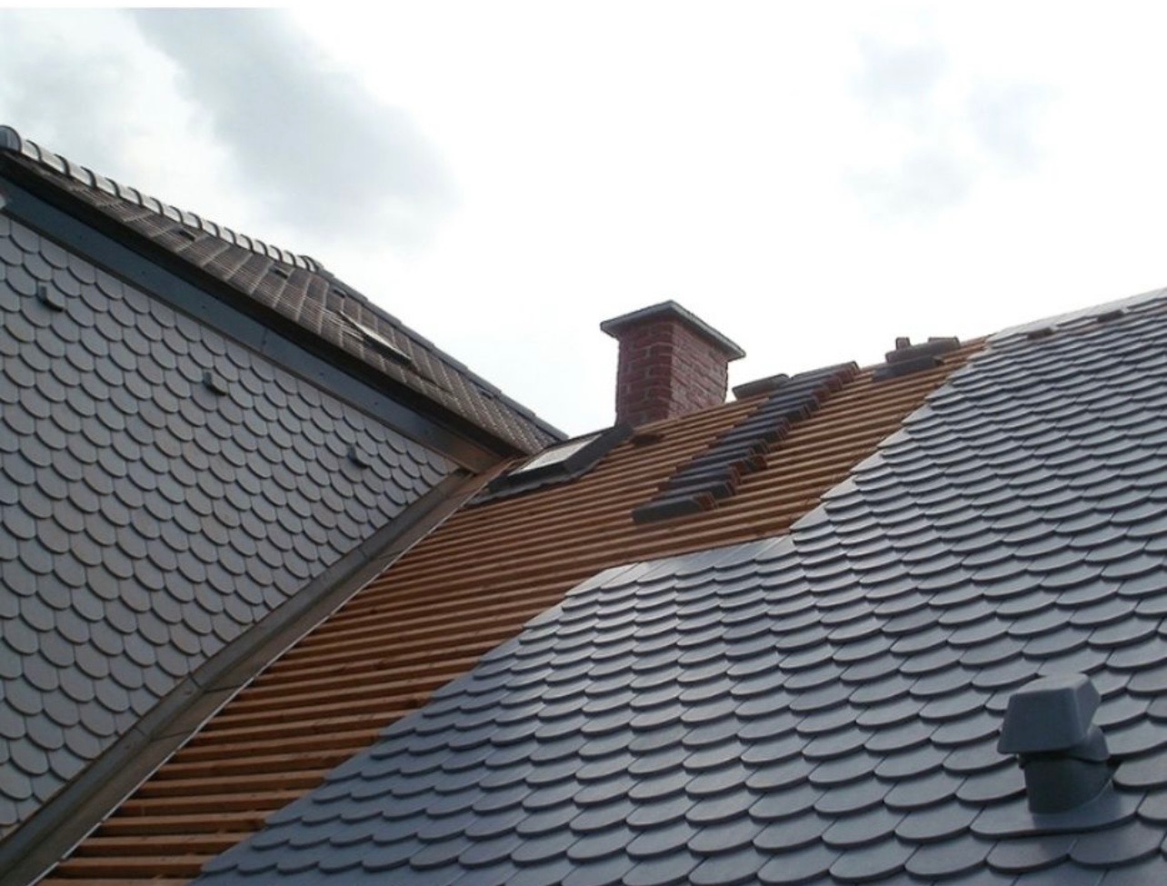 Fragment dachu pokrytego łuską bitumiczną, z widocznym kominem i fragmentem dachu w trakcie układania, ukazującym drewnianą konstrukcję.