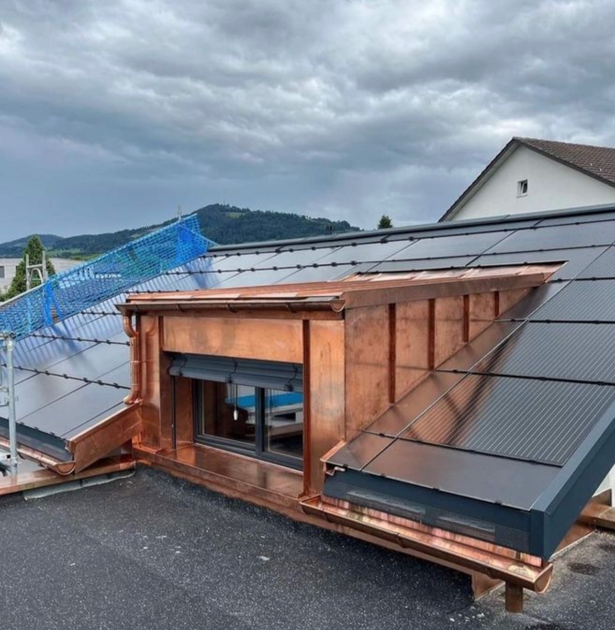 Dach z panelami słonecznymi i miedzianymi elementami wykończeniowymi, w tym obudowa okna dachowego i rynny, na tle zachmurzonego nieba i wzgórz.