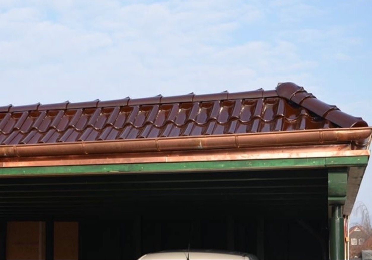 Błyszczący, brązowy dach z ceramicznej dachówki karpiówki, miedziana rynna na zielonej podbitce, widok z dołu na tle błękitnego nieba.