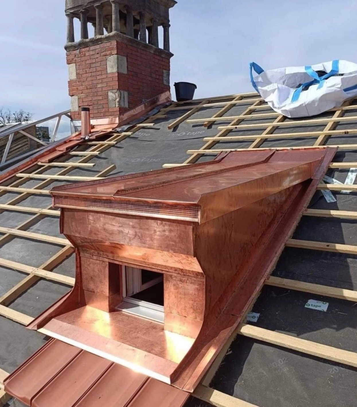 Miedziana lukarna z oknem w trakcie montażu na dachu z drewnianą konstrukcją, widoczny komin z cegły i czarny kubeł.