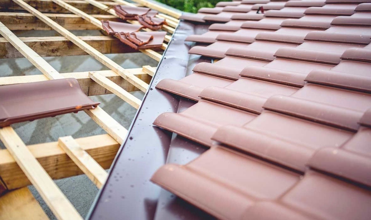 Fragment dachu z drewnianą konstrukcją i brązową dachówką karpiówką w trakcie montażu, widoczne obróbki blacharskie.