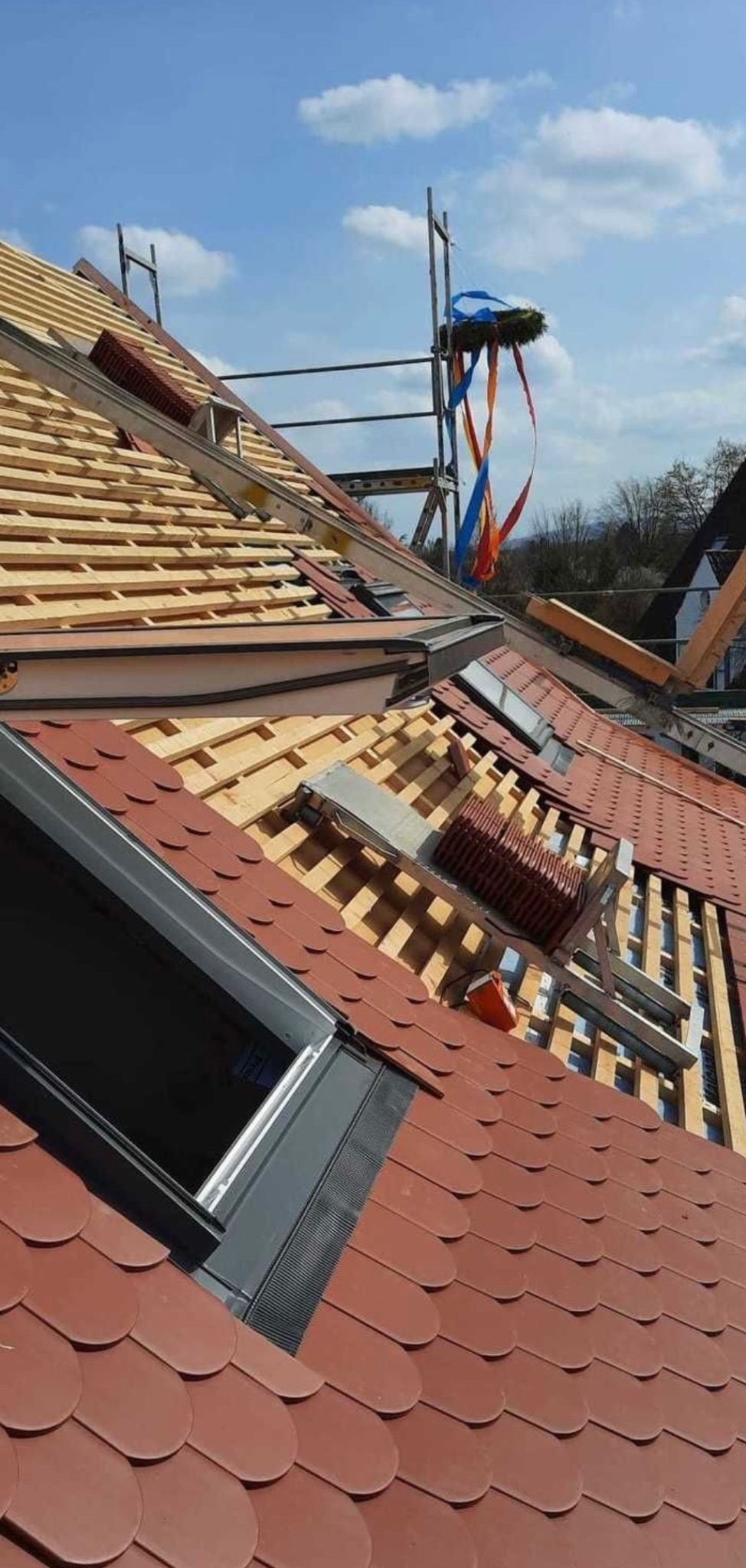 Częściowy widok dachu w trakcie prac z widocznym oknem dachowym, drewnianą konstrukcją, czerwonymi dachówkami i wieńcem na rusztowaniu w tle.