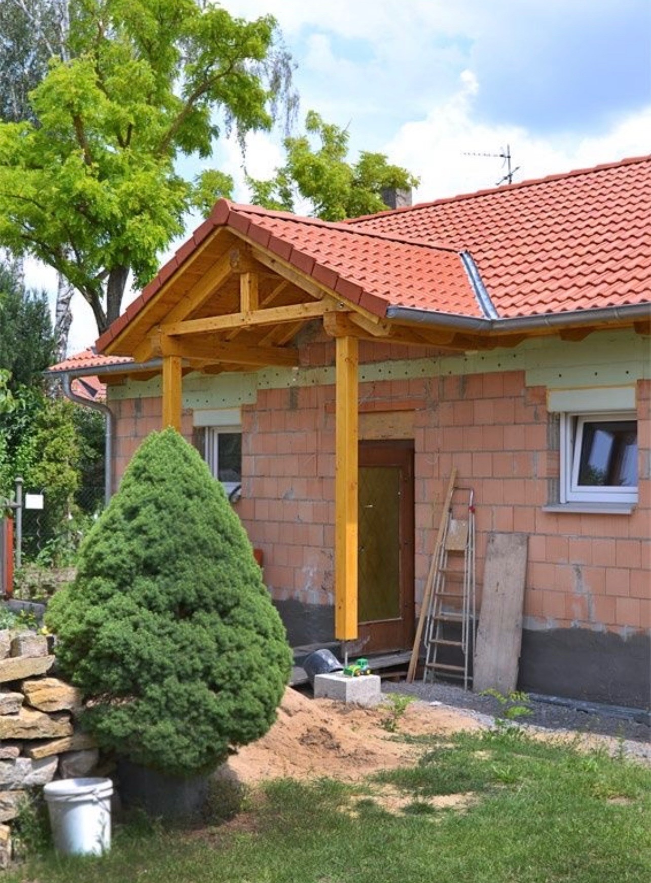 Niedokończona budowa domu z czerwonym dachem, drewnianym zadaszeniem nad wejściem, widoczne rynny, ściany z czerwonej cegły oraz drabina oparta o ścianę.