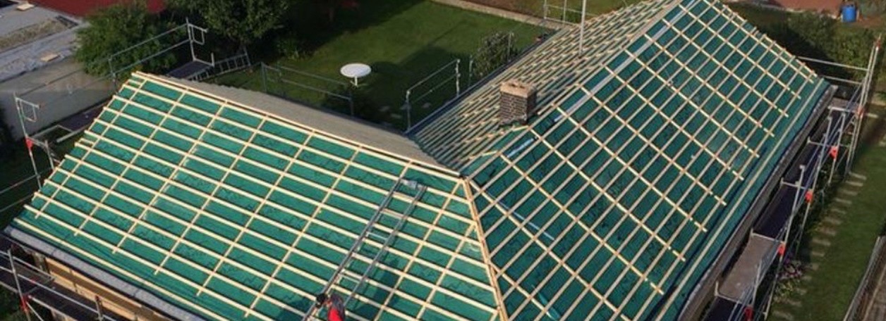 Widok z góry na dach domu w trakcie krycia z zieloną membraną i drewnianą łatą, widoczne rusztowania i komin z cegły.