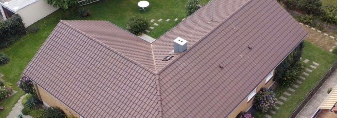 Widok z góry na dwuspadowy dach pokryty brązową dachówką ceramiczną z kominem, otoczony zielonym trawnikiem i ścieżką z płyt.