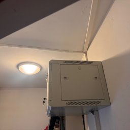 MAŁGORZATA SOBOCIŃSKA - Szara skrzynka alarmowa zamontowana na ścianie wewnątrz budynku w Warszawie. Widoczny fragment sufitu i oświetlenie.