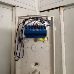 MAŁGORZATA SOBOCIŃSKA - Otwarta obudowa alarmu z niebieską listwą zaciskową, widoczne przewody i poziomica. Biała obudowa z tworzywa przymocowana do ściany, widoczne ślady montażu.