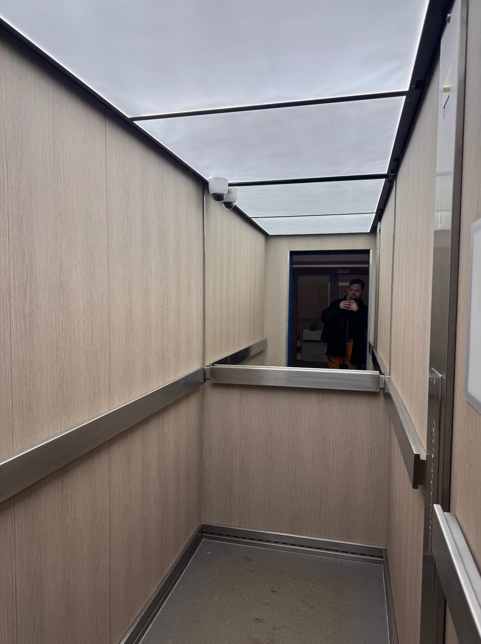 Wnętrze windy wykończonej drewnopodobnymi panelami, z widocznym lustrem, w którym odbija się osoba robiąca zdjęcie. Zamontowane kamery monitoringu w rogu kabiny.