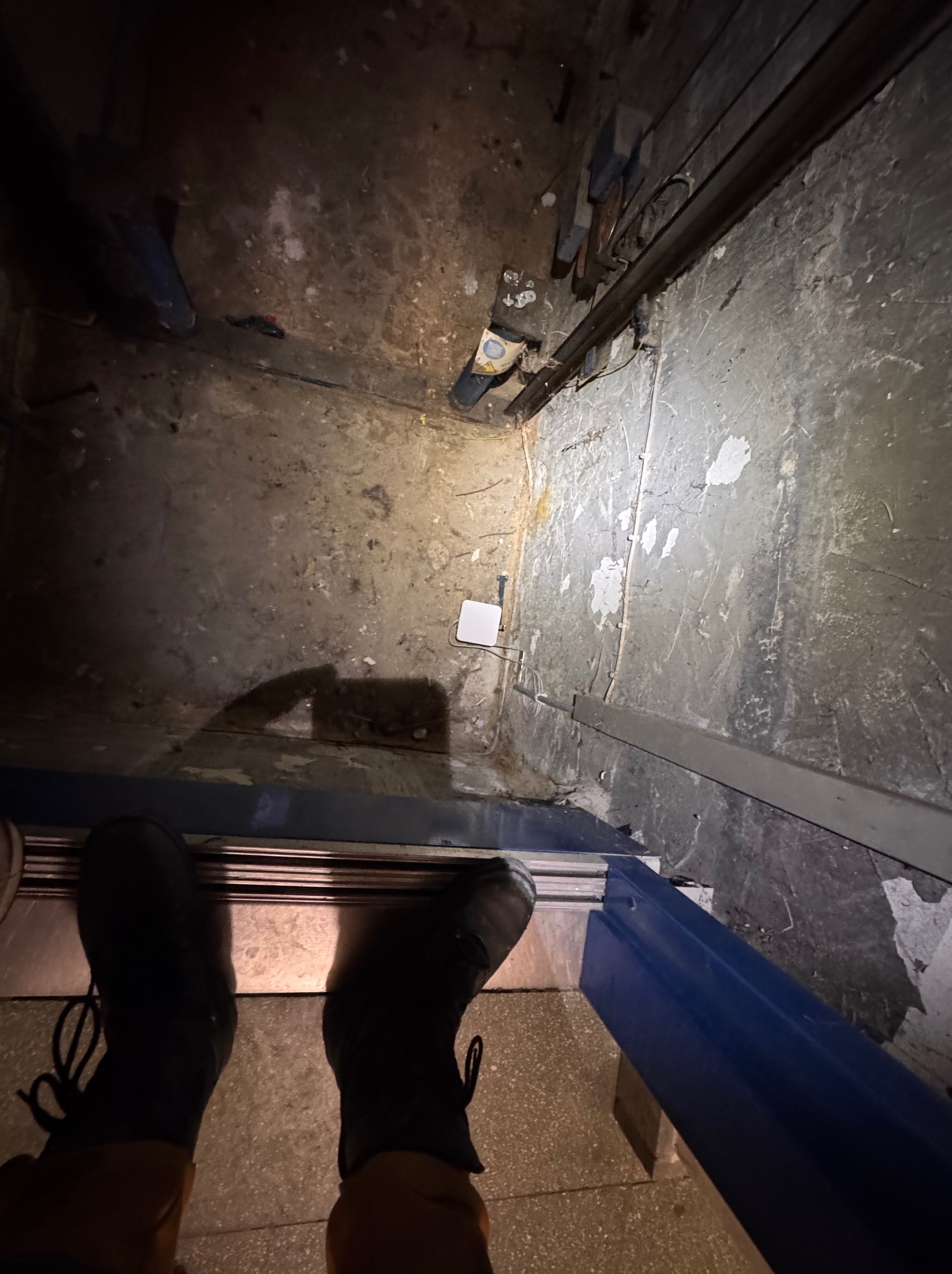 Wnętrze szybu windowego w trakcie prac remontowych. Widoczne elementy konstrukcyjne, ściany z ubytkami tynku i instalacje elektryczne. U dołu kadru fragment butów osoby wykonującej prace.