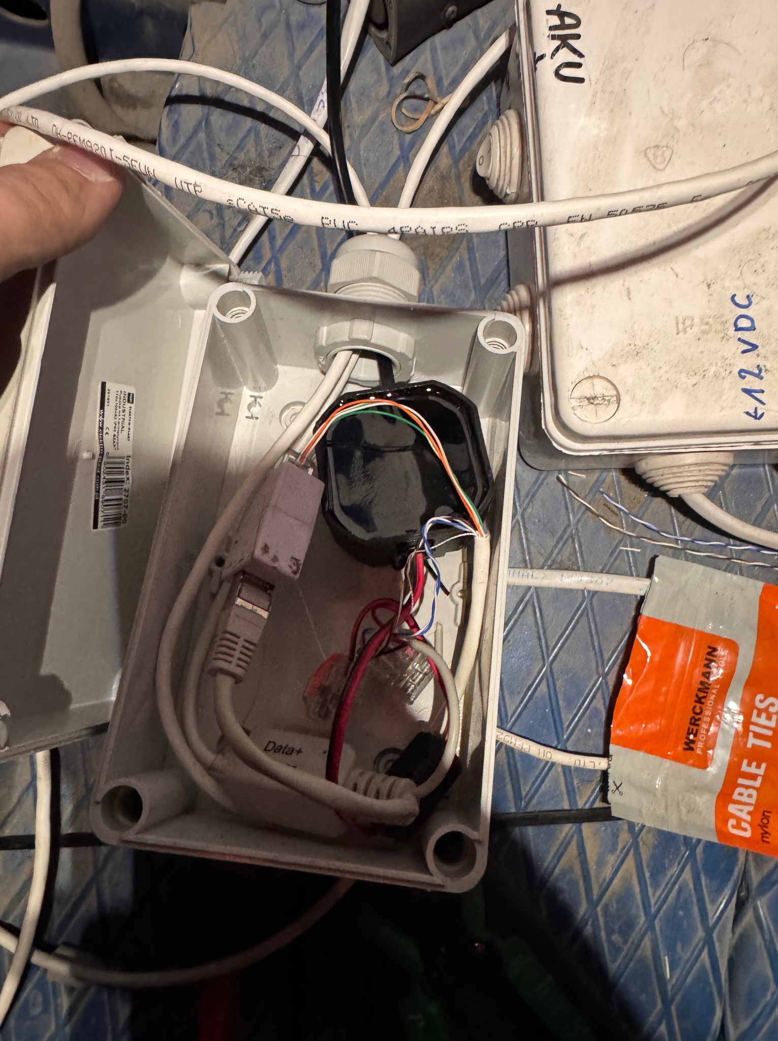 Otwarta puszka instalacyjna z kablami, złączami RJ45 i kolorowymi przewodami. Obok leży opakowanie opasek zaciskowych. Widoczne oznaczenia na kablach i puszkach.