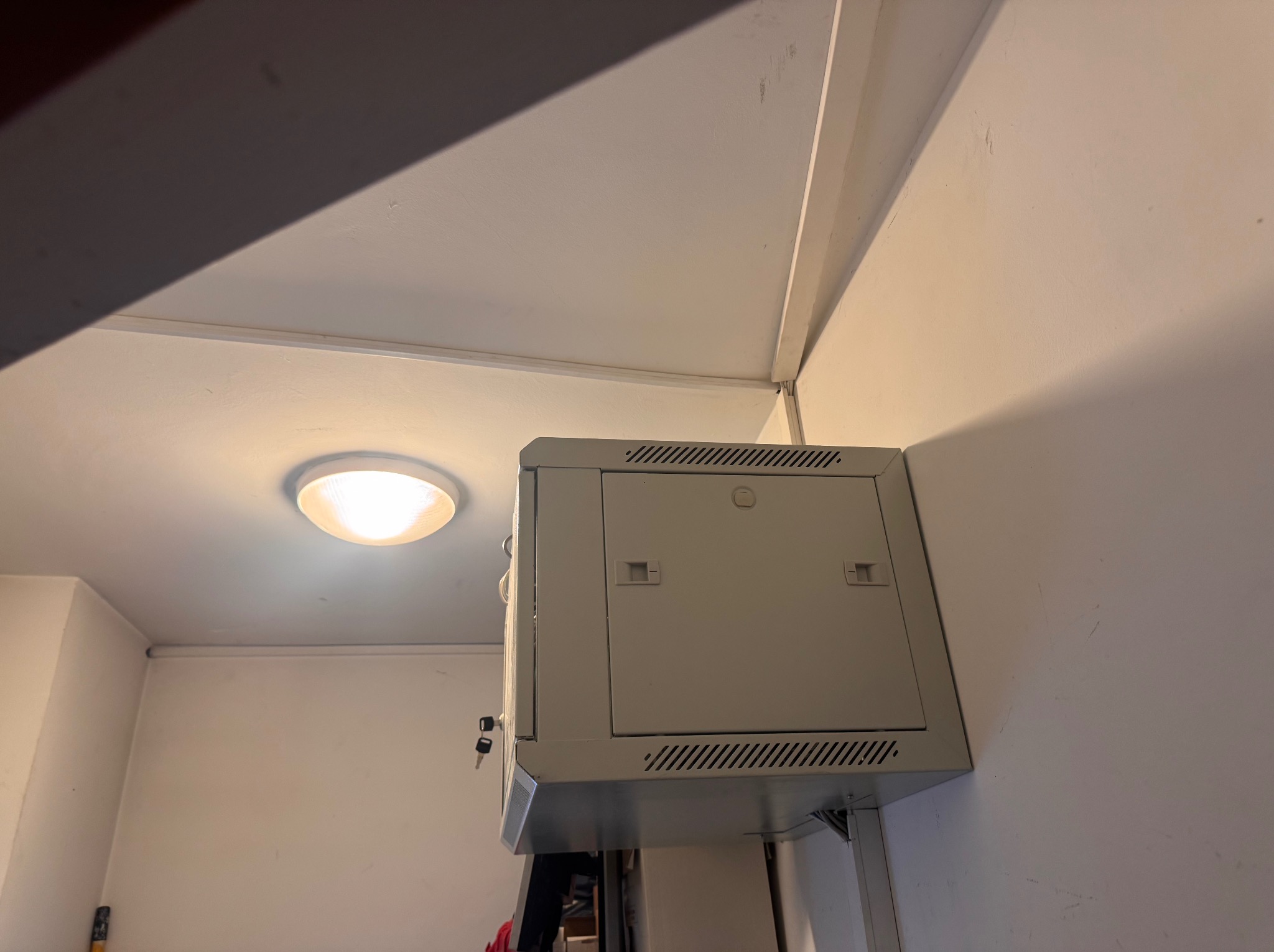 Szara skrzynka alarmowa zamontowana na ścianie wewnątrz budynku w Warszawie. Widoczny fragment sufitu i oświetlenie.