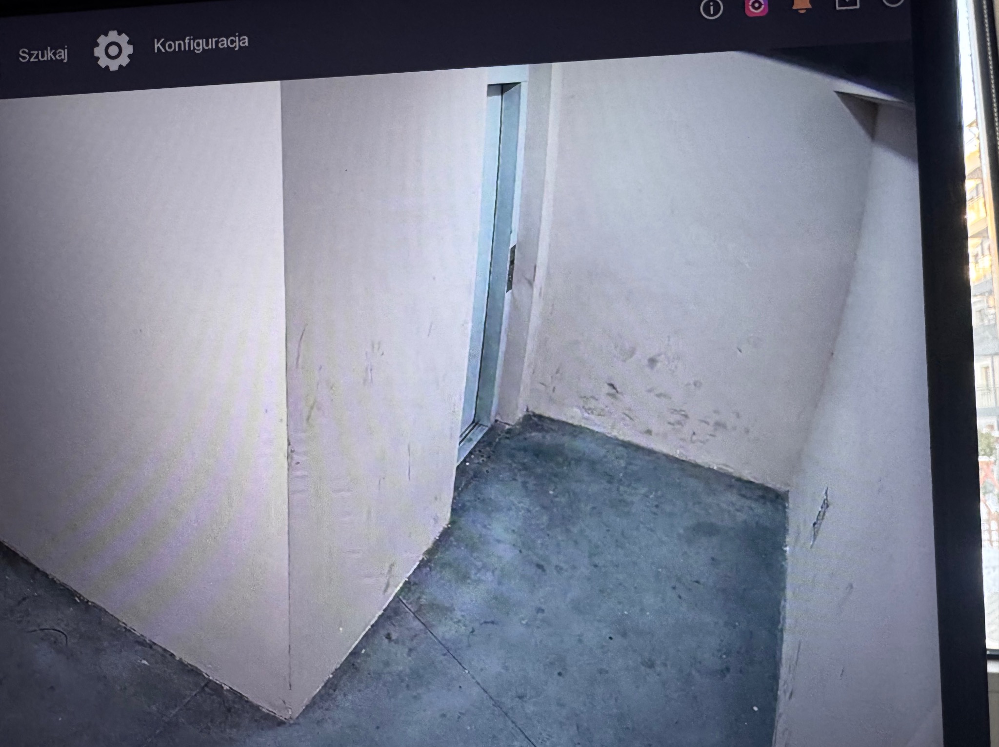 Obraz z kamery monitoringu: surowe wnętrze z widocznymi śladami wilgoci na ścianach i podłodze. W tle widoczne okno i fragment budynku.
