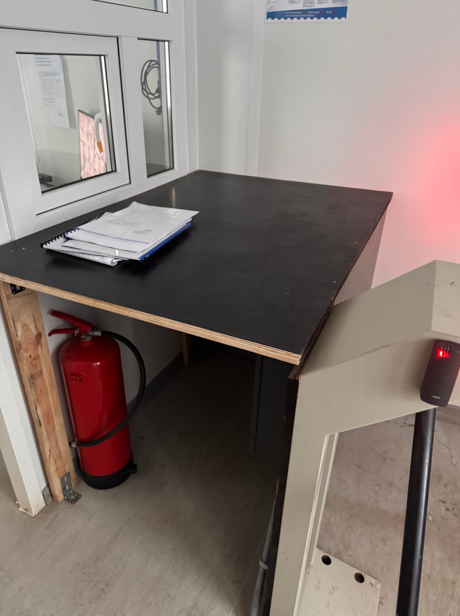 Biurko z dokumentami i gaśnicą w rogu, obok bramka kontrolna z czytnikiem kart. Widoczne okno i instrukcja BHP na ścianie. Oświetlenie mieszane.