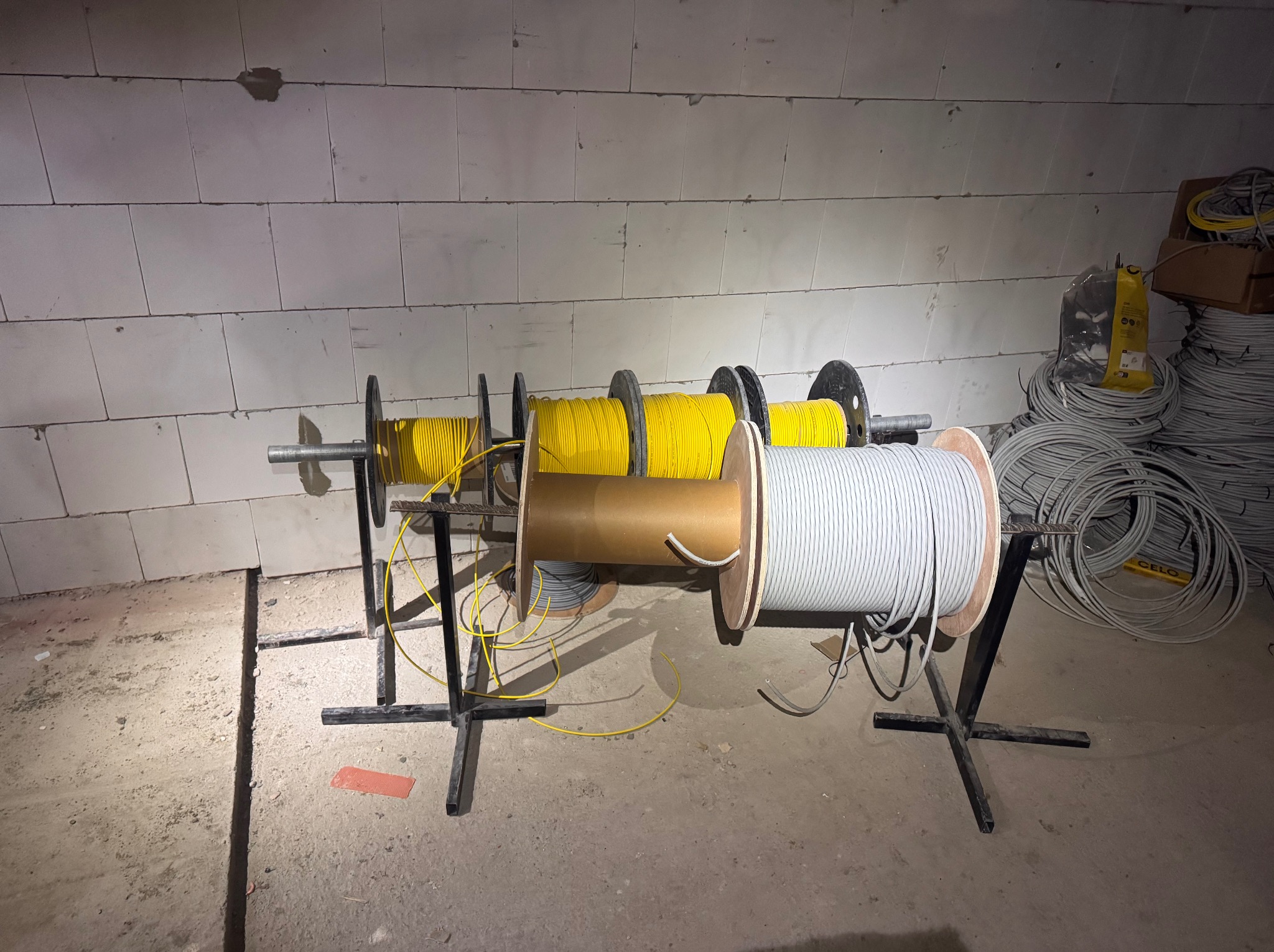 Szkieletowa konstrukcja z rolkami kabli: żółte, szare i brązowe, na tle surowej ściany z betonu komórkowego w trakcie instalacji elektrycznej.