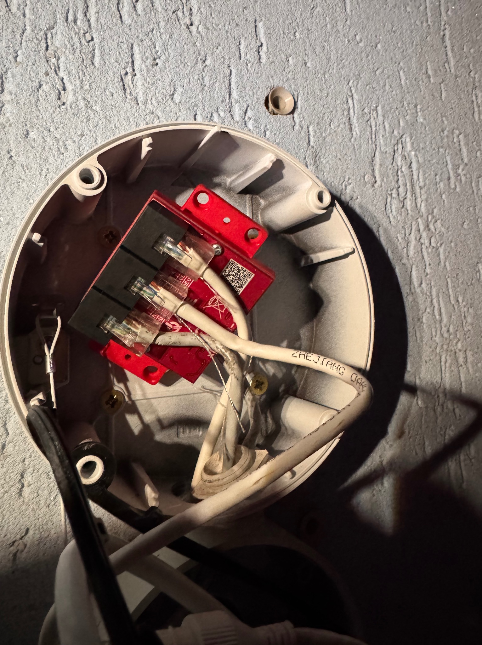 Wnętrze puszki instalacyjnej z czerwonym modułem alarmowym, widoczne przewody i złącza. Montaż alarmu z bliska na szarej, teksturowanej ścianie.