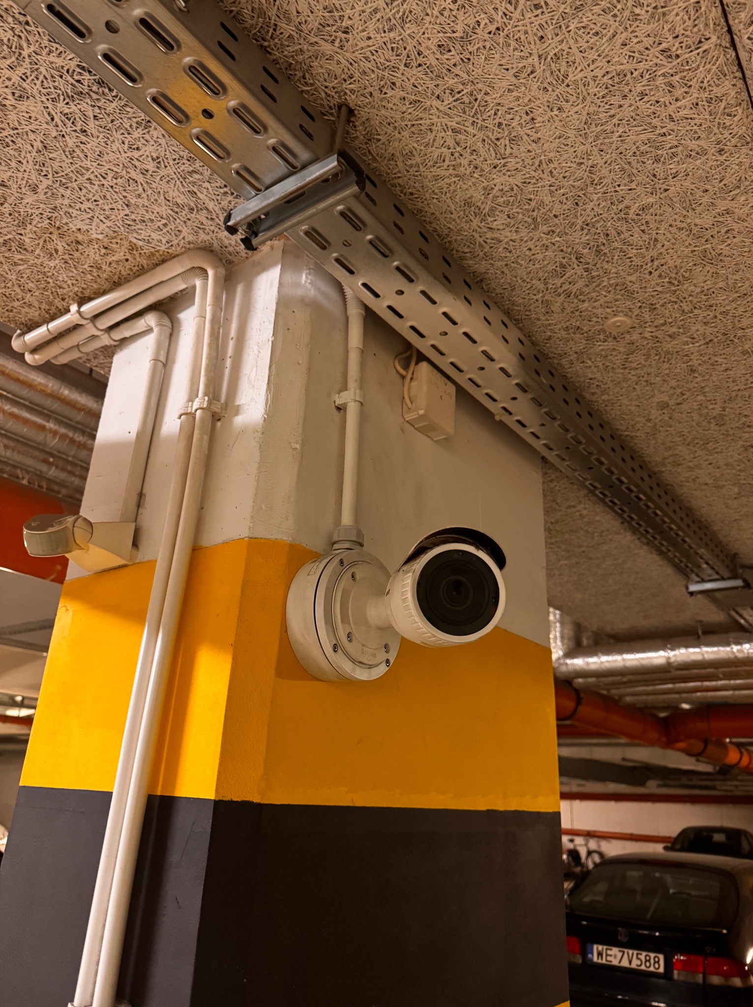 Kamera monitoringu zamontowana na kolumnie w garażu podziemnym, widoczne rury instalacyjne i fragment zaparkowanego samochodu. Oświetlenie sufitowe.