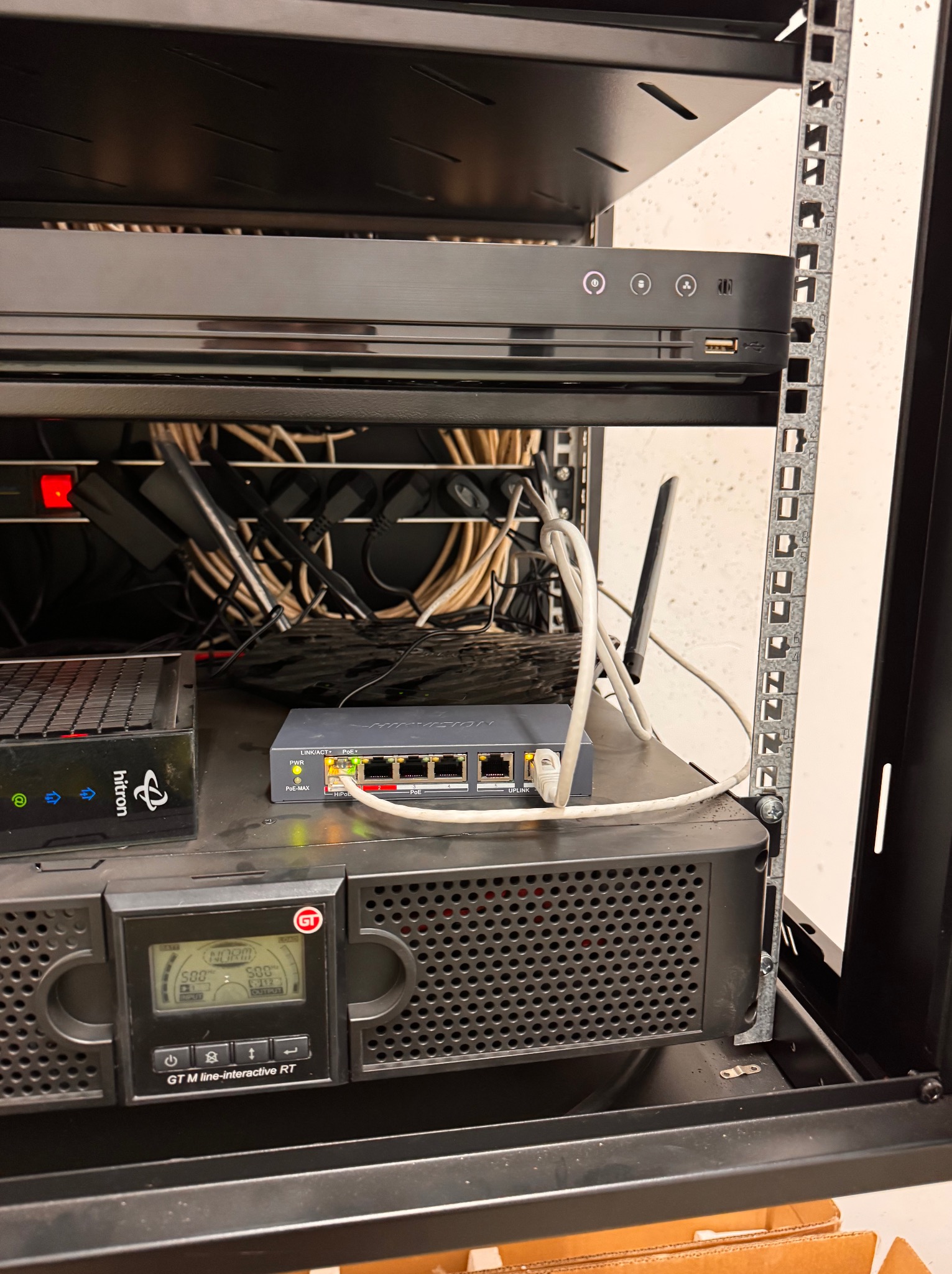Szafa rack z urządzeniami sieciowymi: router, switch, UPS. Widoczne kable, zasilacze i elementy montażowe. Sprzęt HiTron i GT M line-interactive RT.