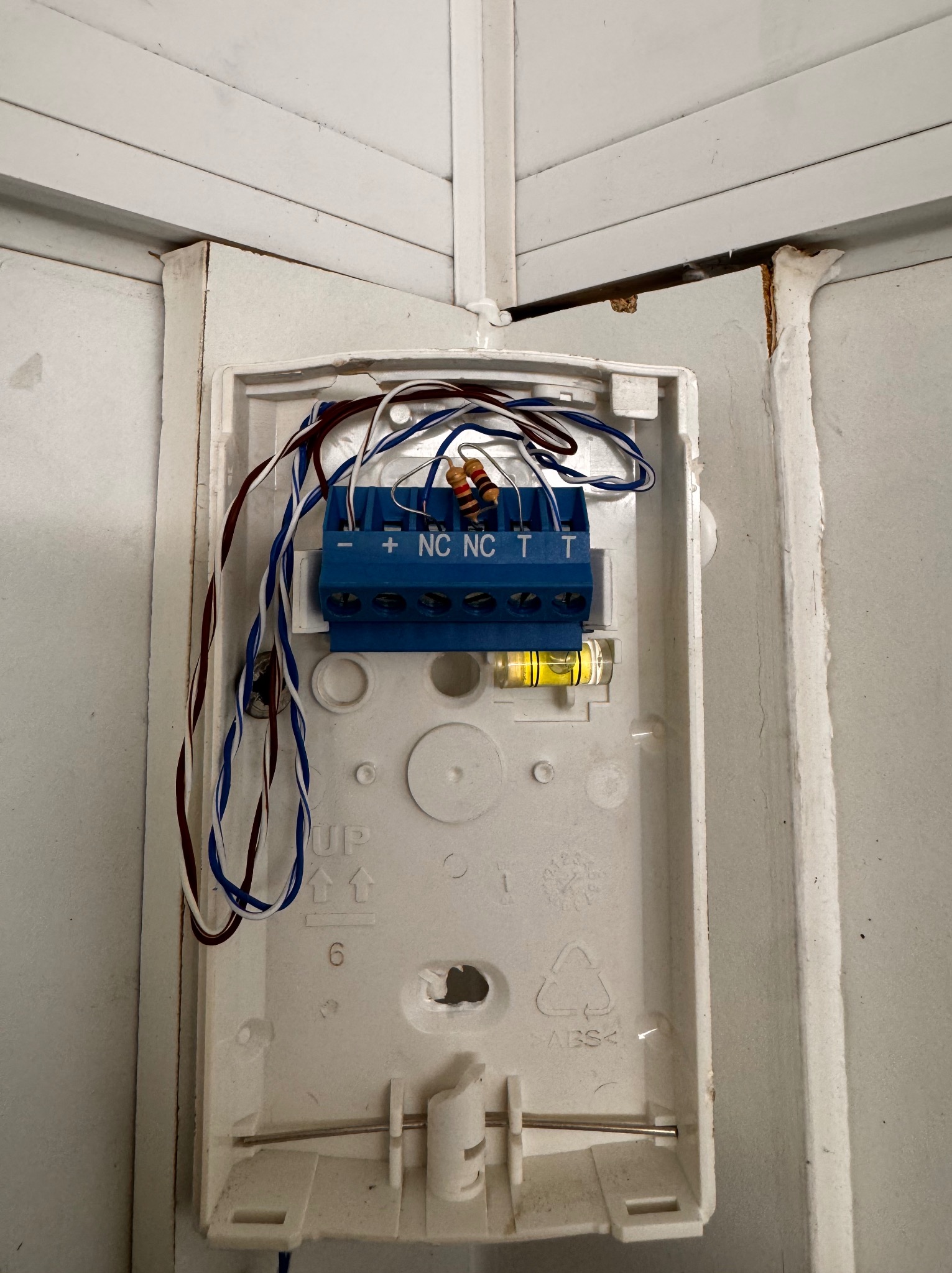 Otwarta obudowa alarmu z niebieską listwą zaciskową, widoczne przewody i poziomica. Biała obudowa z tworzywa przymocowana do ściany, widoczne ślady montażu.