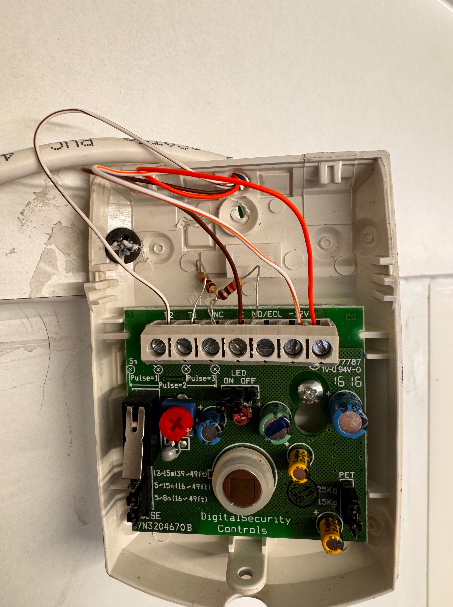 Otwarta obudowa czujnika alarmowego z widoczną płytką drukowaną, przewodami i elementami elektronicznymi. Widoczne oznaczenia i napisy na płytce.