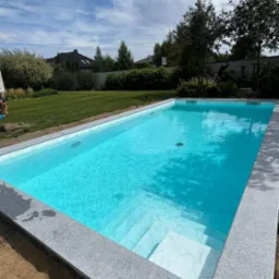 Nowoczesny basen z szarym, kamiennym obramowaniem i białymi stopniami, otoczony trawnikiem i drzewami, w słoneczny dzień.