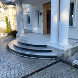 Wejście do domu z kolumnami i trzema stopniami z ciemnego kamienia, wyłożone kostką granitową w odcieniach szarości z widocznym odpływem liniowym.